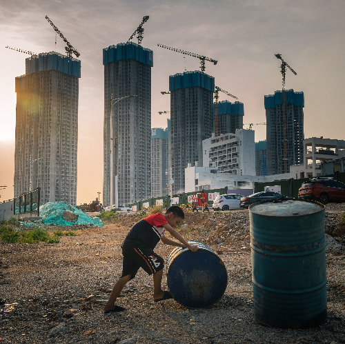 在深圳市光明区，一个小孩正在推着油桶玩耍，后面的高楼大厦正拔地而起。.jpg